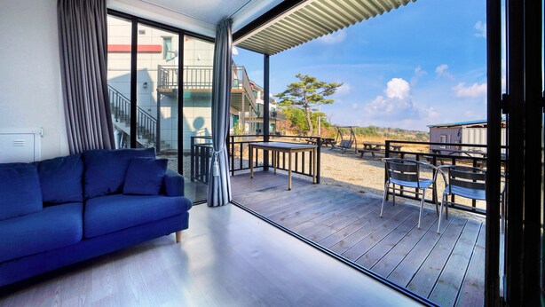 A modern living room with a blue sofa, a glass door leading to an outdoor deck, and a clear view of the beach and buildings in the background. - Gyeongju Hyu Plus Pension (Gyeongju)