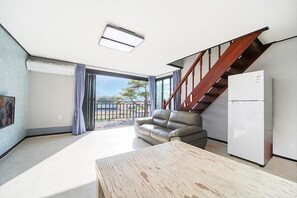 Una sala de estar moderna con una mesa de madera, un sofá gris, un refrigerador blanco y una escalera que lleva a un balcón.