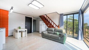 A modern living room with a grey sofa, a wooden staircase, and large windows offering a view of trees. - Gyeongju Hyu Plus Pension (Gyeongju)