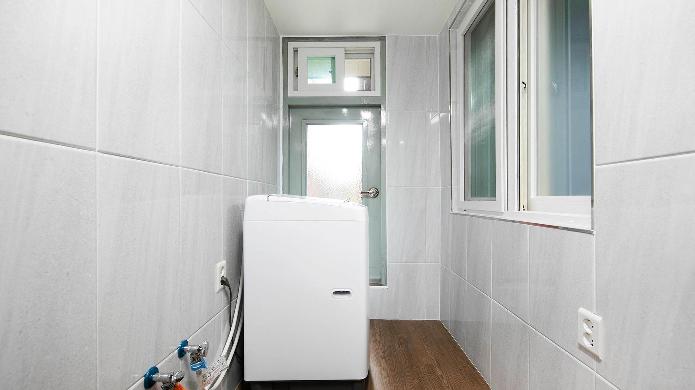 A narrow hallway with white tiled walls, a white refrigerator, a window with a view, and a door with a small window above it.
