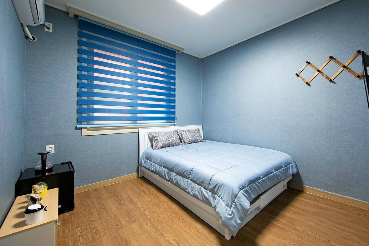 Et soveværelse med seng, et vindue med rullegardiner, trægulv, et lille bord med lampe og et ophængt fjernsyn.