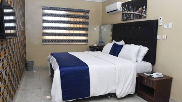 Een hotelkamer met een bed, een nachtkastje, een wandairconditioner, een raam met jaloezieën en een stadsgezicht schilderij.