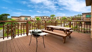 Une terrasse en bois avec un barbecue et une table de pique-nique, entourée d’une clôture en bois et bordée, au loin, de bâtiments en briques.