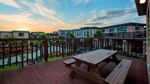 Une terrasse en bois, agrémentée de tables de pique-nique, offre une vue sur un quartier résidentiel avec des bâtiments en briques et une rivière.