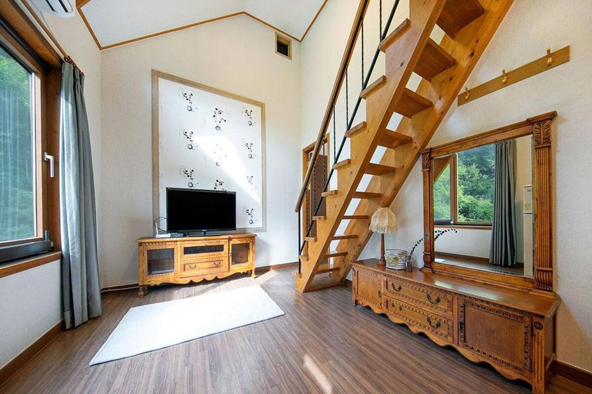 Une pièce avec un escalier en bois, un grand miroir, un téléviseur posé sur un meuble en bois et une fenêtre avec des rideaux.