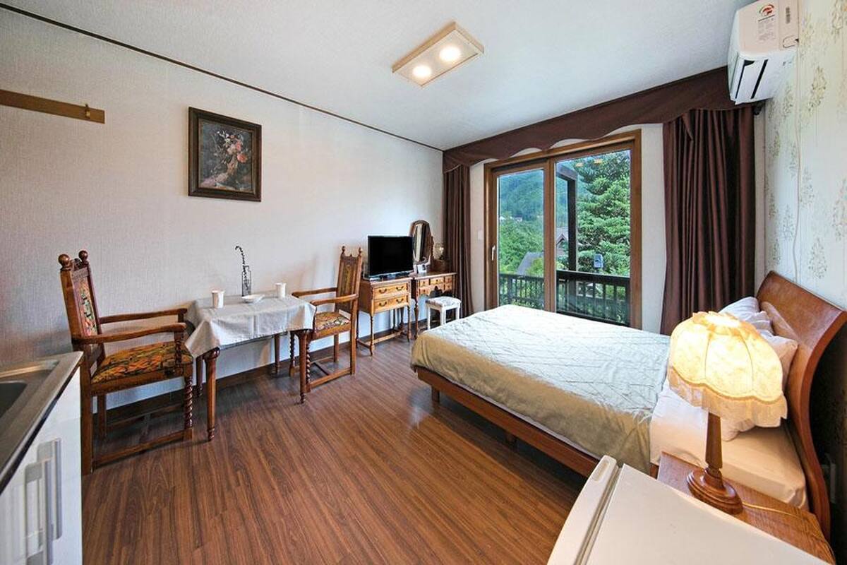 Une chambre d’hôtel équipée d’un lit, d’un bureau, d’une chaise, d’une télévision et d’une fenêtre donnant sur des arbres.