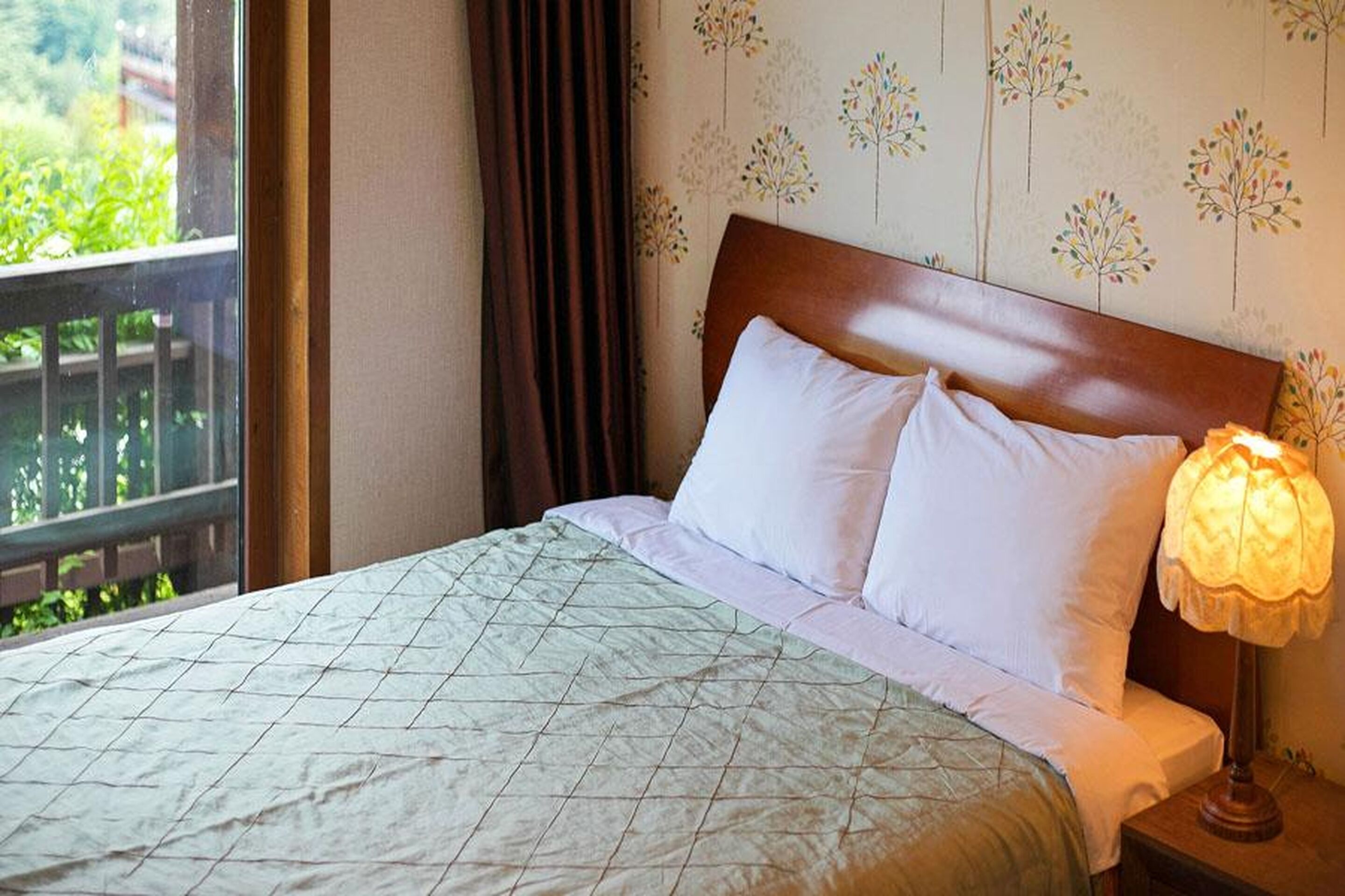 A hotel room with a bed, a nightstand, a lamp, and a window with a view of greenery.