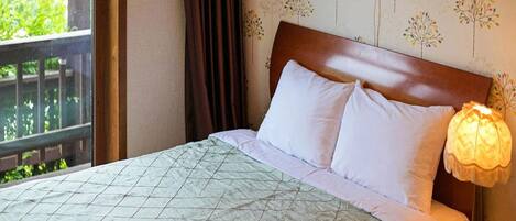 A hotel room with a bed, a nightstand, a lamp, and a window with a view of greenery.