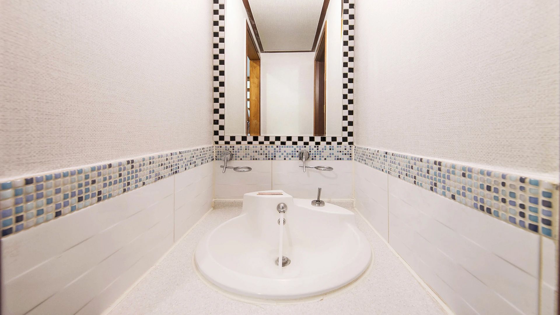 A bathroom with a white sink, two faucets, and a mirror above. The walls have a checkered tile pattern near the floor.