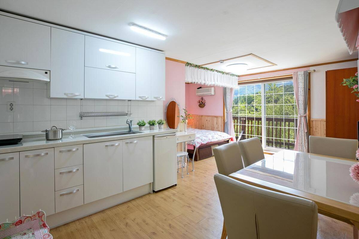 Une cuisine compacte avec des meubles blancs, un coin repas doté d’une table en verre et une chambre à coucher avec un lit et une fenêtre.