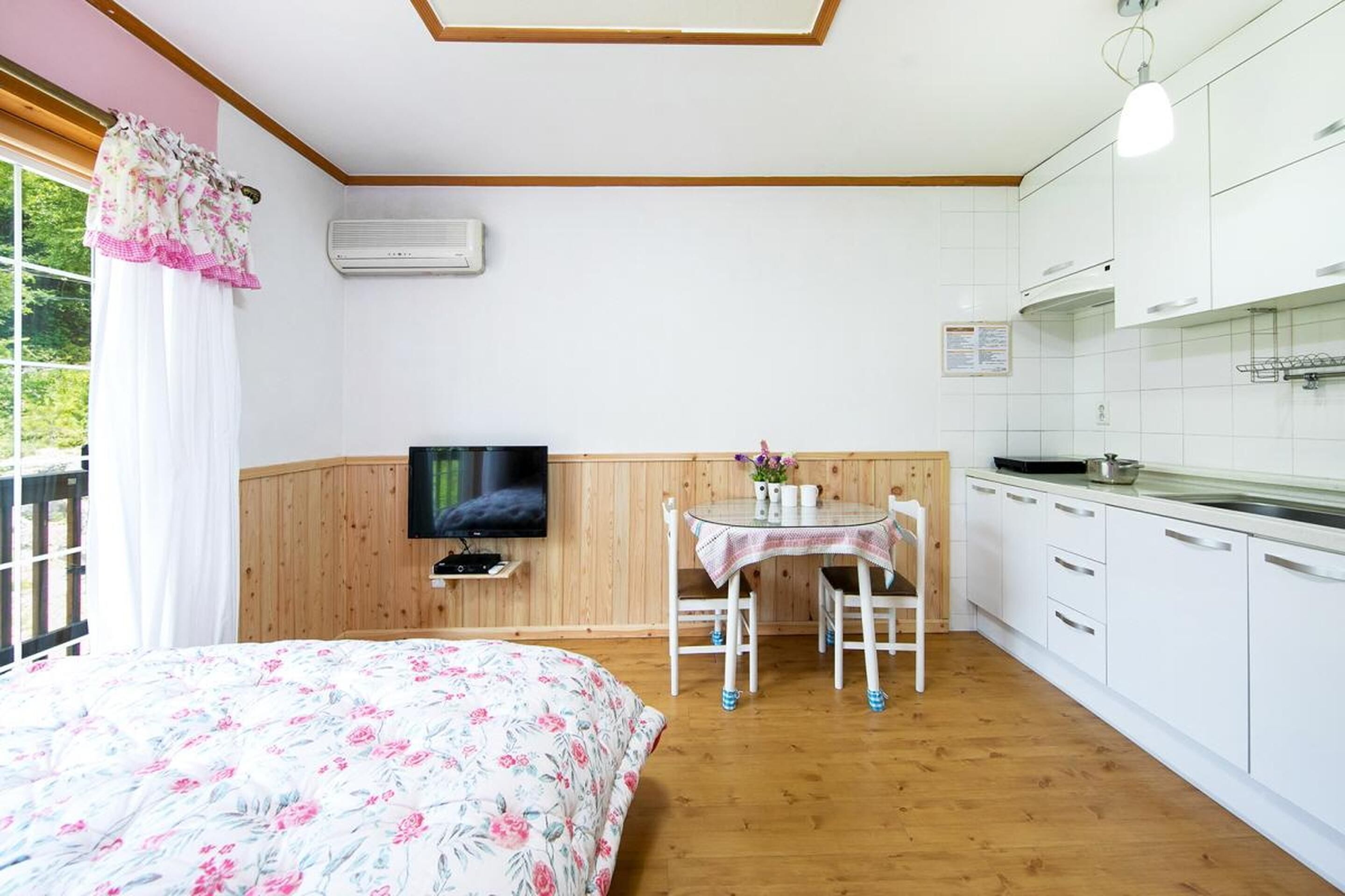 A bedroom with a floral bedspread, a television on a wooden shelf, a small table with chairs, and a kitchen area with white cabinets and a wooden countertop.