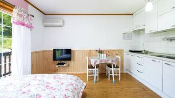 A bedroom with a floral bedspread, a television on a wooden shelf, a small table with chairs, and a kitchen area with white cabinets and a wooden countertop.