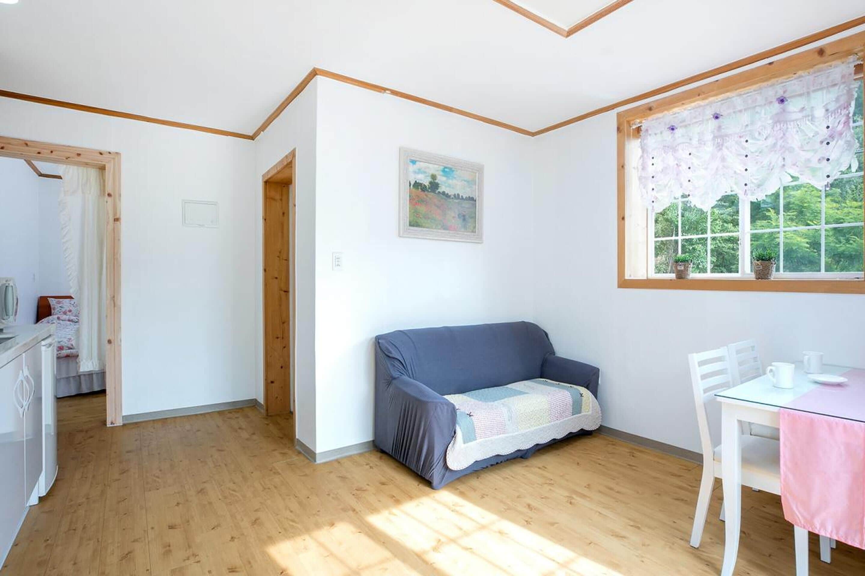 A room with a wooden floor, a blue sofa, a white table with a pink tablecloth, and a window with a floral curtain.