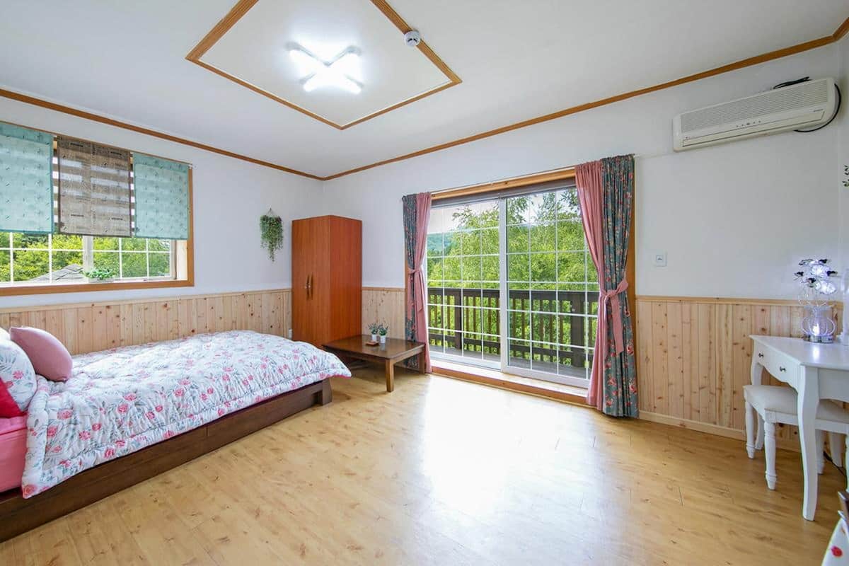 Une chambre avec un plancher en bois, un lit recouvert d’une literie à motifs floraux, un bureau et une fenêtre avec des rideaux.