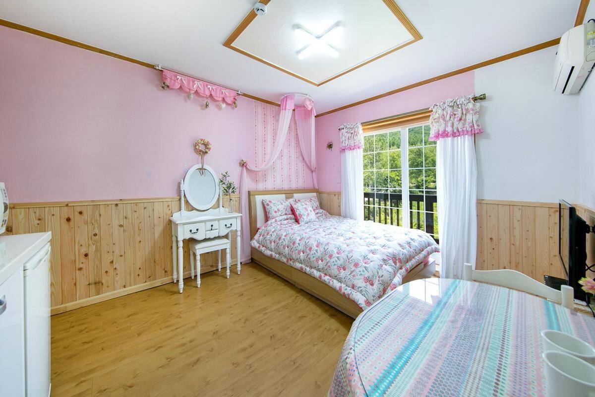 Une chambre avec un plancher en bois, un lit avec une literie à motifs floraux, une commode blanche, une table ronde avec des tasses et une fenêtre avec des rideaux.