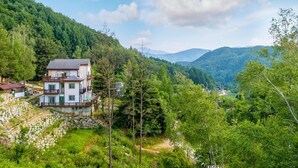 Ein zweistöckiges Haus mit Balkon, umgeben von üppiger Vegetation und Bergen im Hintergrund.