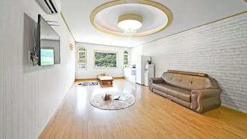 A modern living room with a brown leather sofa, a flat-screen TV mounted on the wall, and a kitchen area with white appliances.