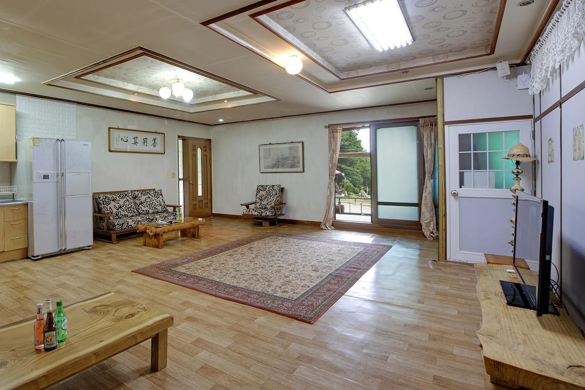 A spacious living area with a wooden floor, a sofa, a coffee table, and a large rug. There is a kitchen area with cabinets and a refrigerator.