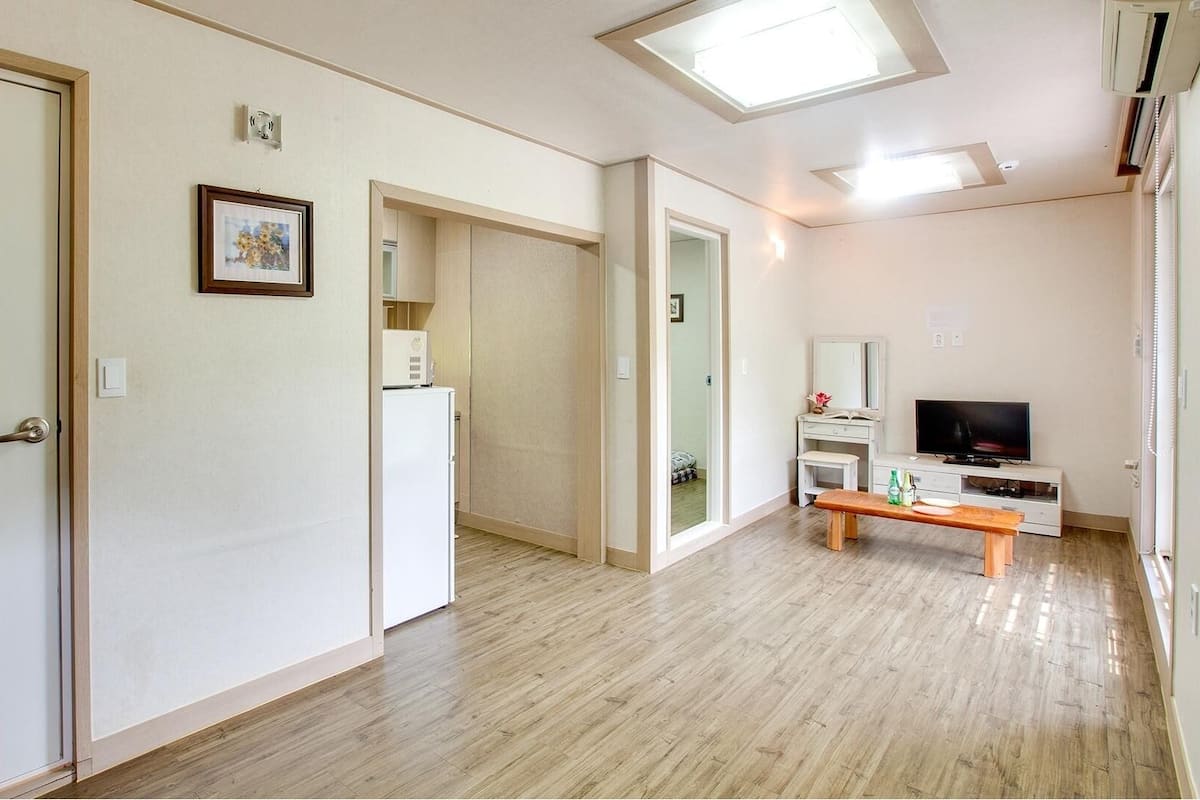 A spacious living area with a wooden floor, a television, a bench, and a refrigerator.