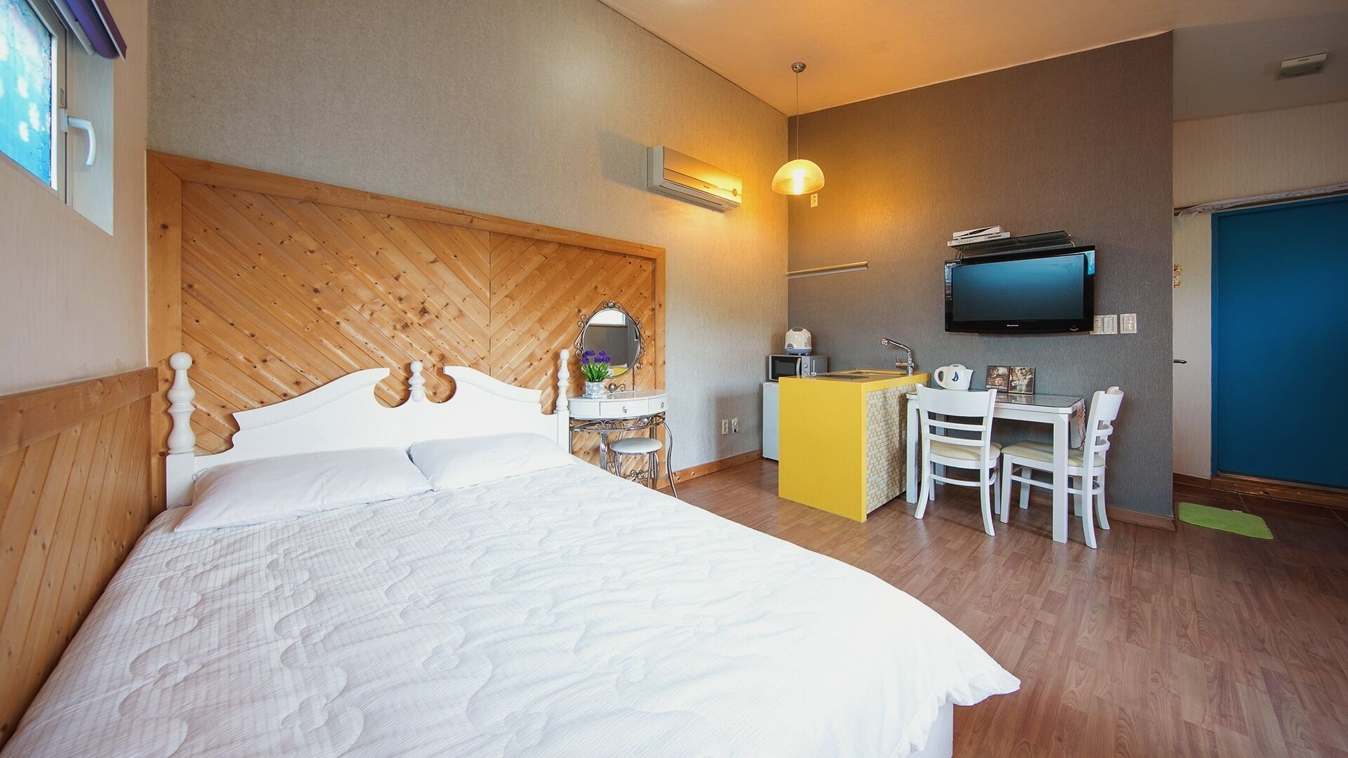 Un dormitorio con cabecera de madera, una cama, un escritorio con silla, un televisor y una kitchenette pequeña.