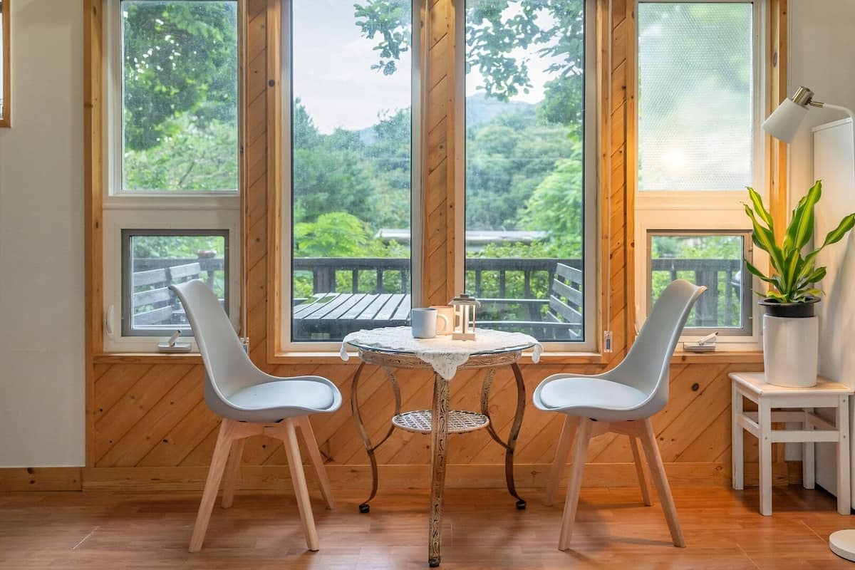 Une piĂšce avec un plancher en bois, deux chaises blanches, une petite table ronde, un rĂ©frigĂ©rateur blanc et de grandes fenĂȘtres donnant sur un paysage verdoyant.