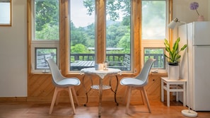 Una habitación con piso de madera, dos sillas blancas, una mesita redonda pequeña, un refrigerador blanco y ventanas amplias con vista a follaje.