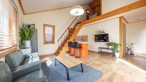Una sala de estar moderna con un sofá, una mesa de centro, una escalera, una zona de cocina y un televisor montado en la pared.