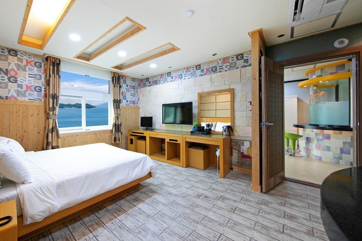 A modern hotel room with a bed, desk, TV, and a view of the sea.