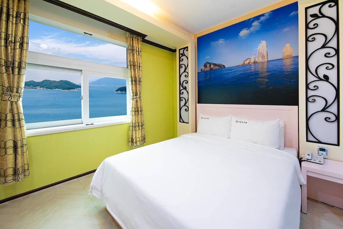 A hotel room with a bed, a nightstand, a painting of a coastal scene, and a window with curtains.