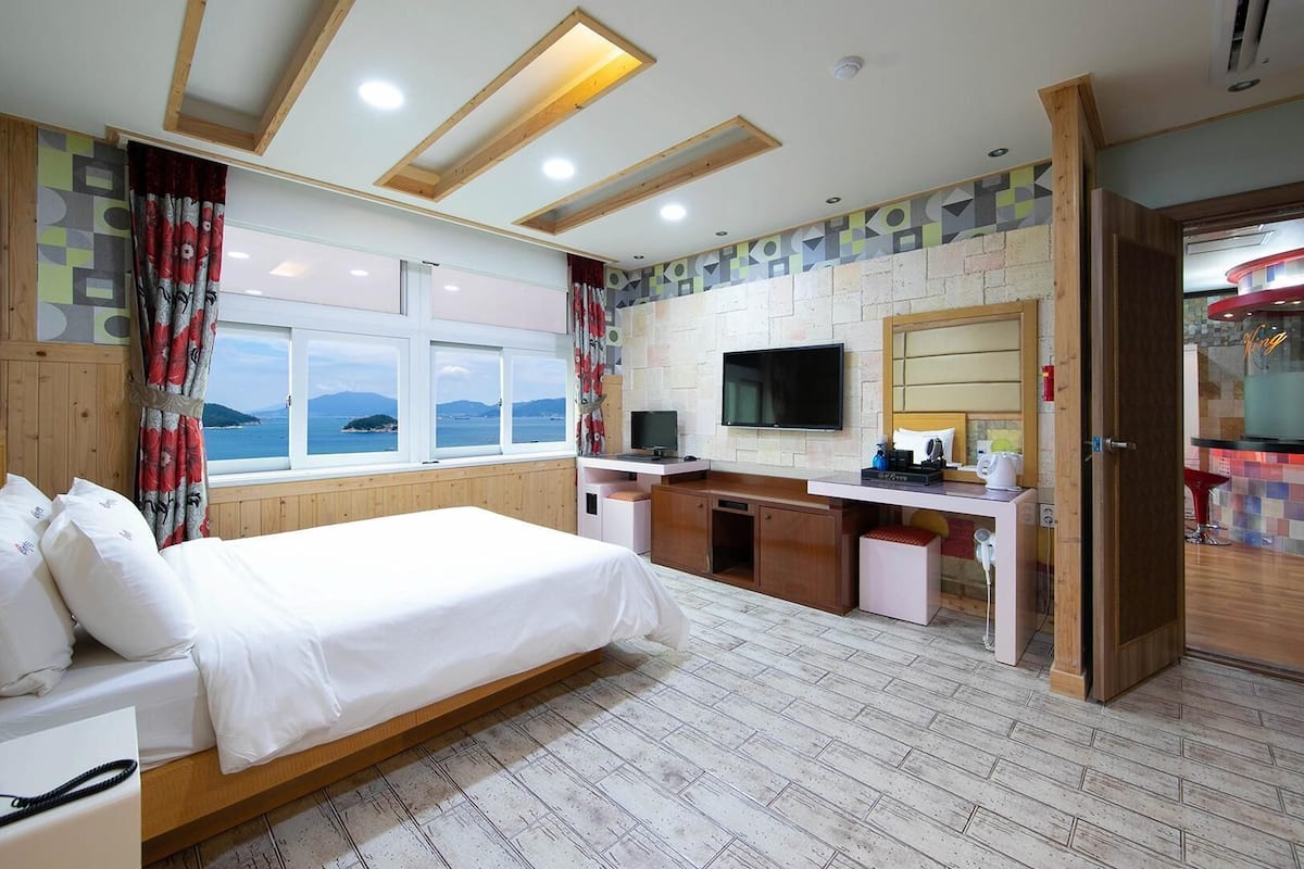 A hotel room with a bed, a desk, a TV, and a view of the sea.