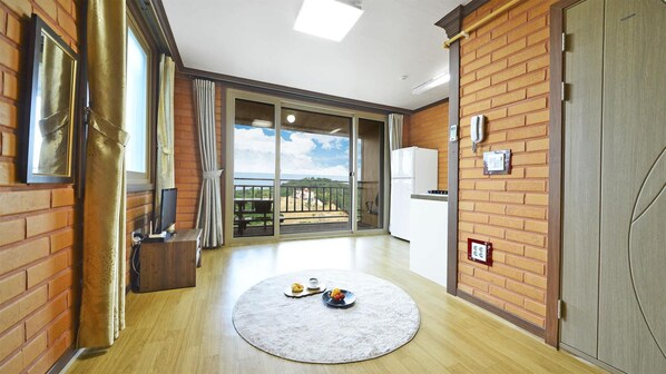 Una habitación amplia con piso de madera, un gran ventanal corredero que da a un balcón con vistas al paisaje, y un tapete redondo pequeño con una cesta de frutas encima.