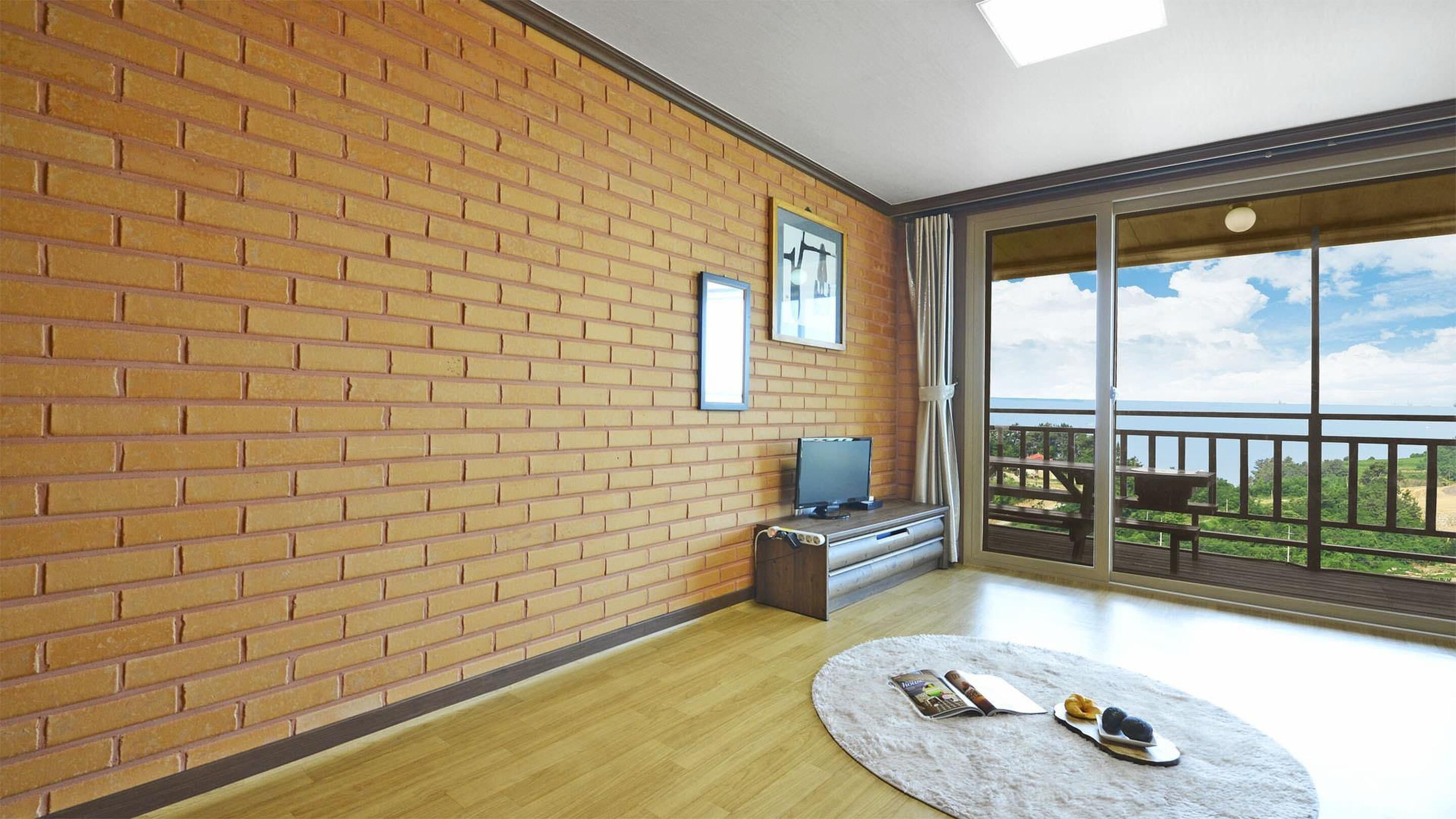 A room with a brick wall, a television on a wooden stand, a round rug with a book and snacks, and a balcony with a view.