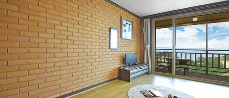 A room with a brick wall, a television on a wooden stand, a round rug with a book and snacks, and a balcony with a view.