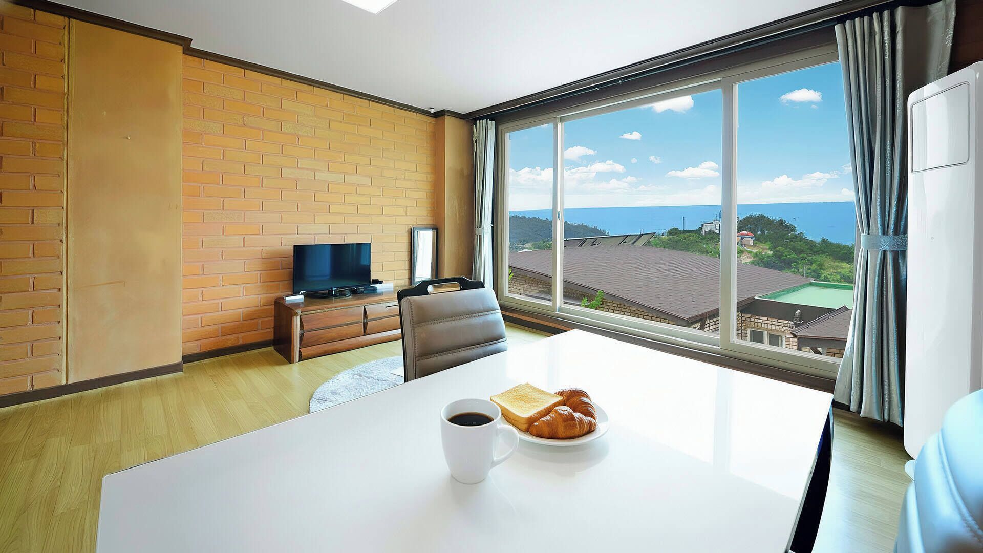 A hotel room with a large window, a flat-screen TV, a dining table with breakfast, and a brick wall.