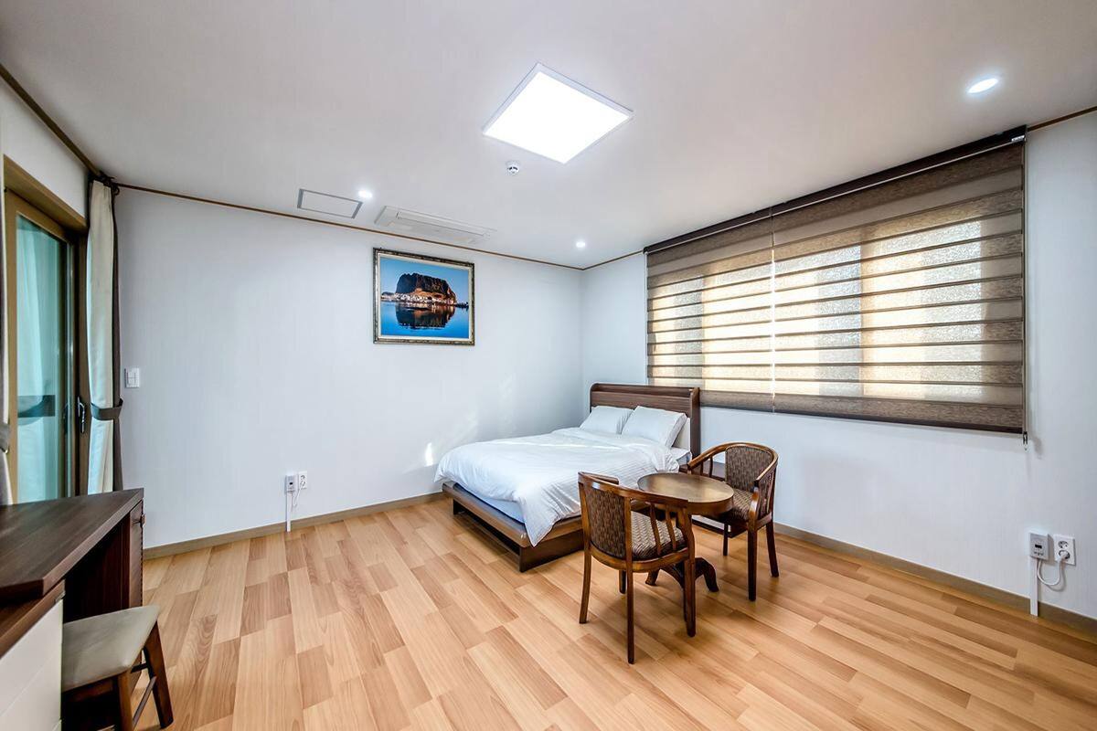 Ein Schlafzimmer mit Bett, Schreibtisch, Stuhl, einem Fenster mit Jalousien und einem Bild an der Wand.