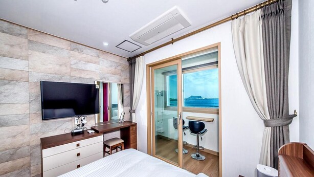 Une chambre d’hôtel avec un lit, un bureau avec une télévision, une chaise et une porte vitrée donnant sur un balcon avec vue sur la mer.