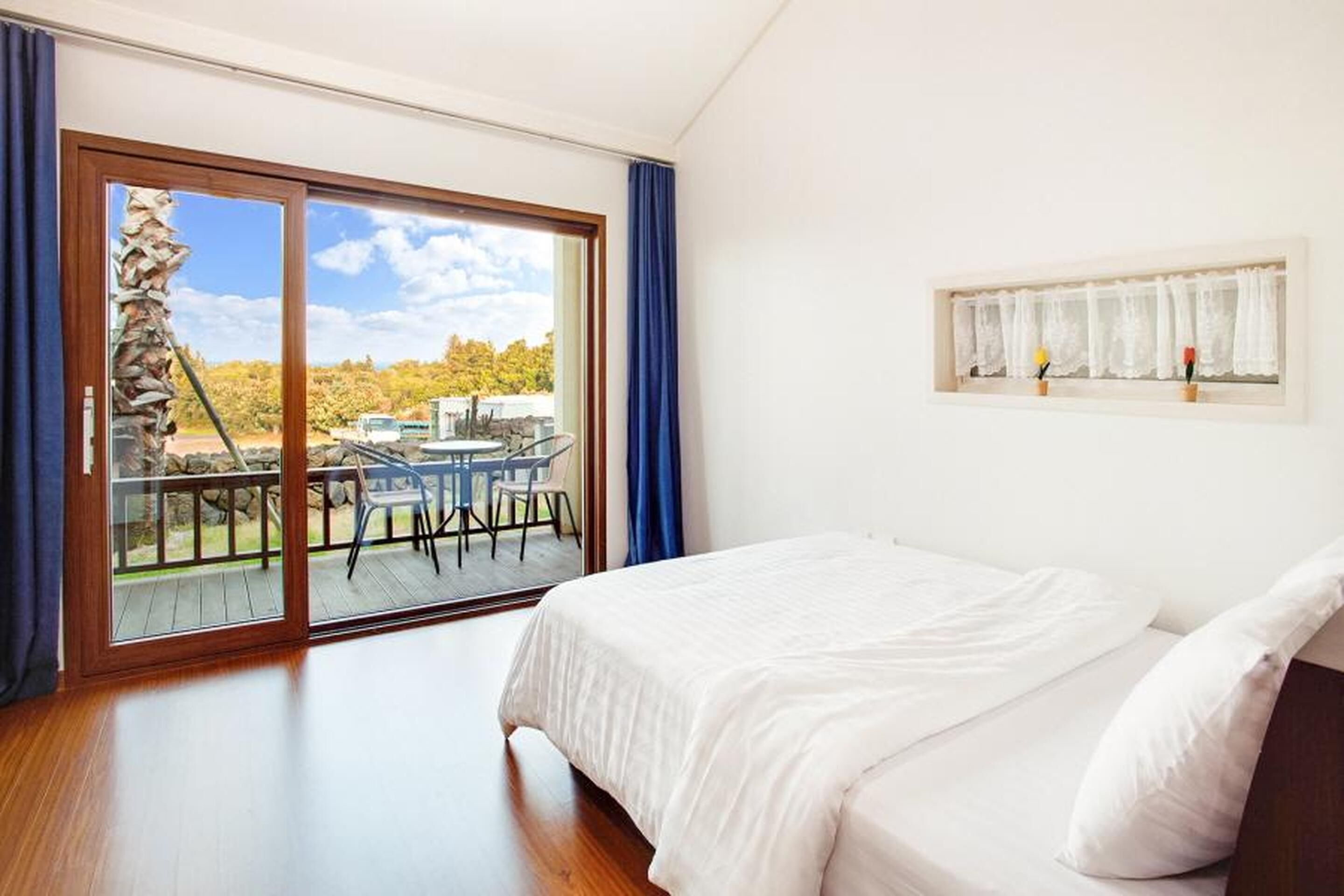A bedroom with a large sliding glass door leading to an outdoor deck, a bed with white linens, and a small window with lace curtains.