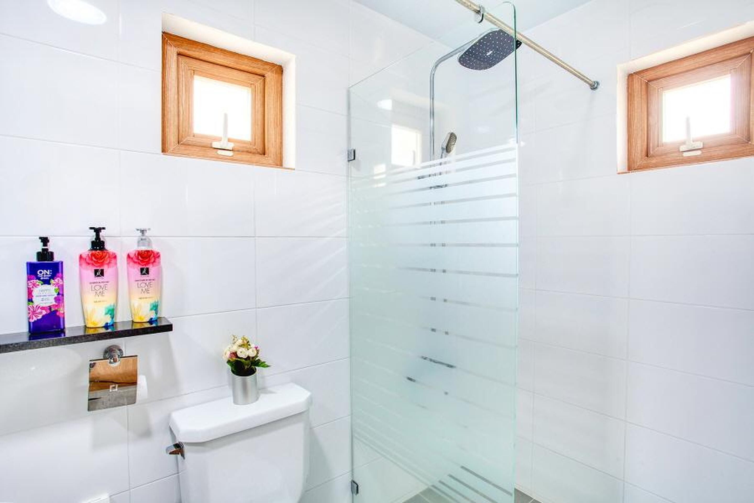 Een moderne badkamer met witte tegels, een glazen douchecabine, een toilet, een wastafel met zeepdispensers en twee kleine ramen.