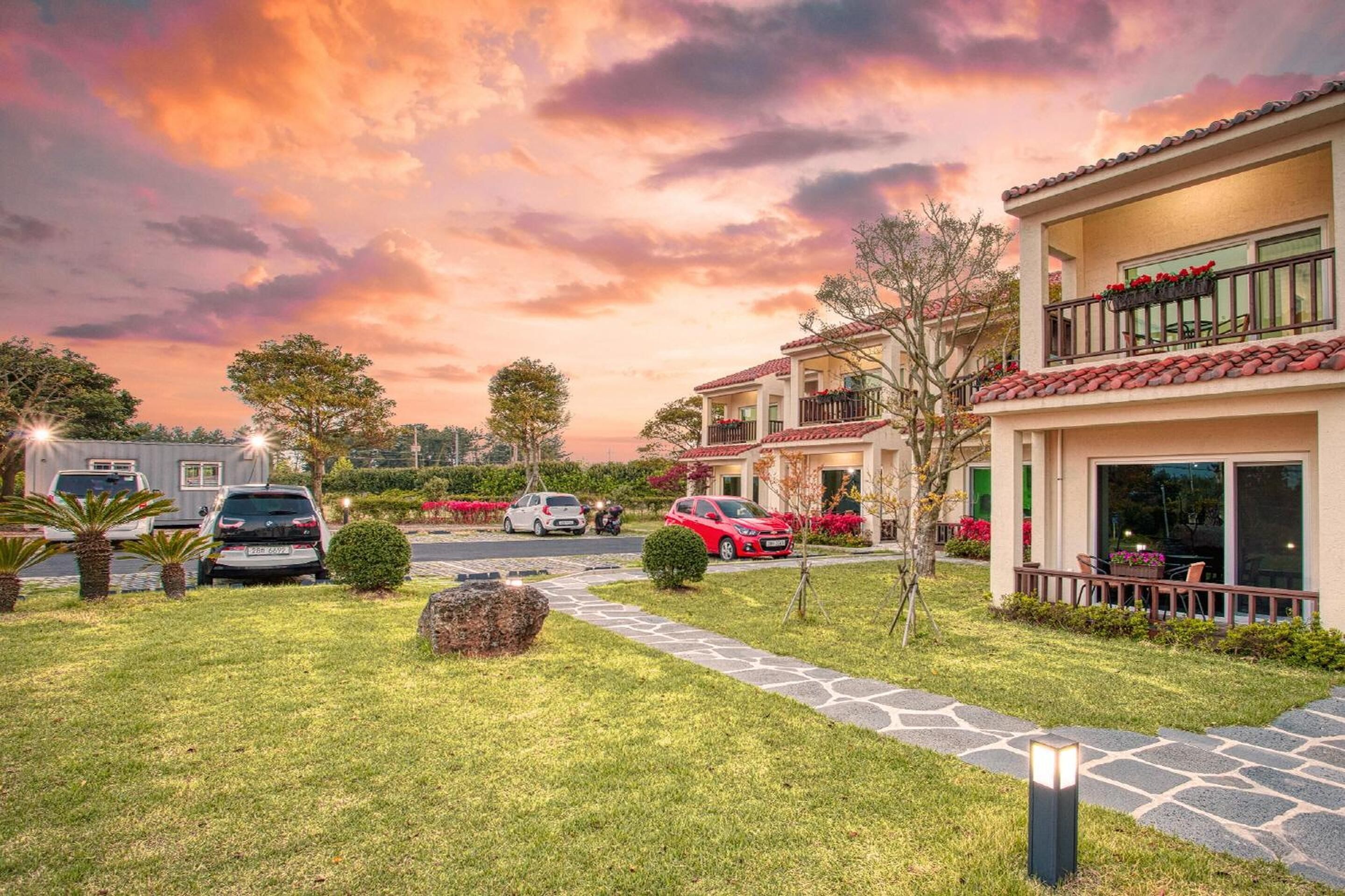 A residential area with a house featuring a balcony with potted plants, a paved driveway, and a well-maintained lawn.