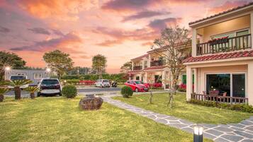A residential area with a house featuring a balcony with potted plants, a paved driveway, and a well-maintained lawn.