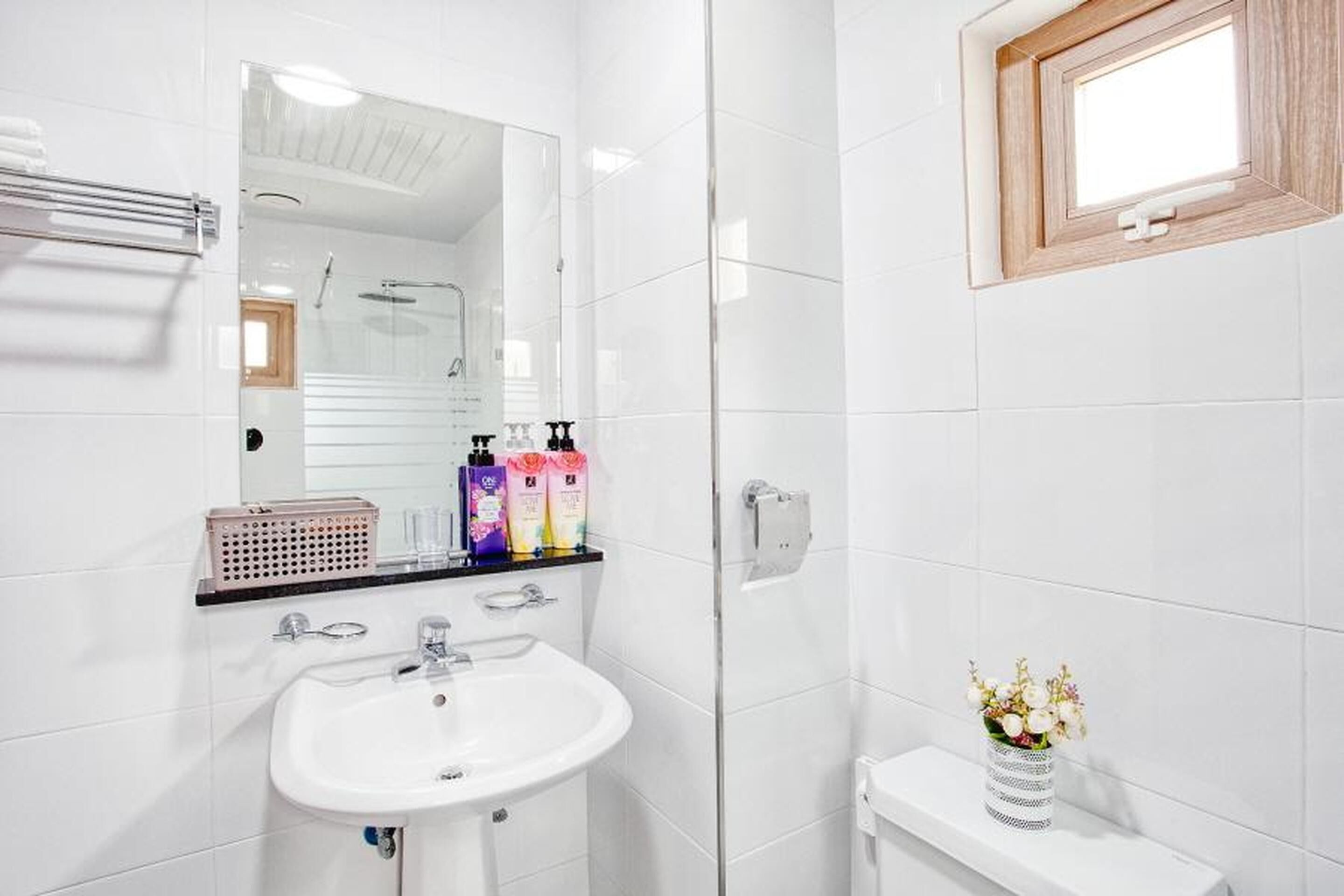 A modern bathroom with a white sink, mirror, and shower. There are toiletries on the counter and a vase with flowers on the sink.