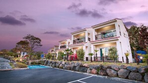 A two-story hotel with a balcony on each floor, a stone retaining wall, and a parking area. - Jeju Kiaora Resort (Seogwipo)