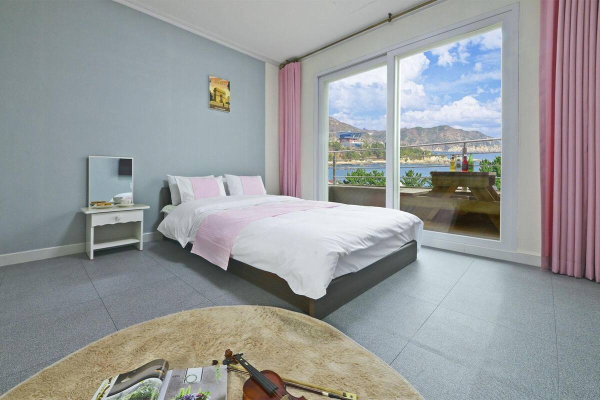 Une chambre à coucher avec un lit, une table de chevet, une fenêtre donnant sur un beau paysage, et une petite table sur laquelle se trouvent un violon et un magazine.