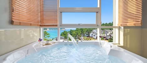 A hot tub with a view of a coastal landscape through a window with wooden blinds.