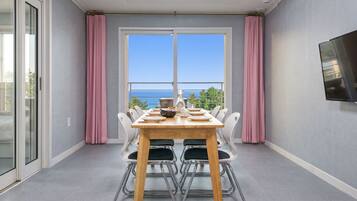 A dining area with a table set for four, a TV mounted on the wall, and a view of the sea through large windows.