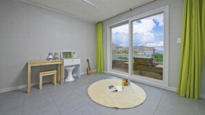 Een kamer met een groot raam, een houten bureau, een witte stoel, een gitaar en een rond vloerkleed met flessen en een tijdschrift.