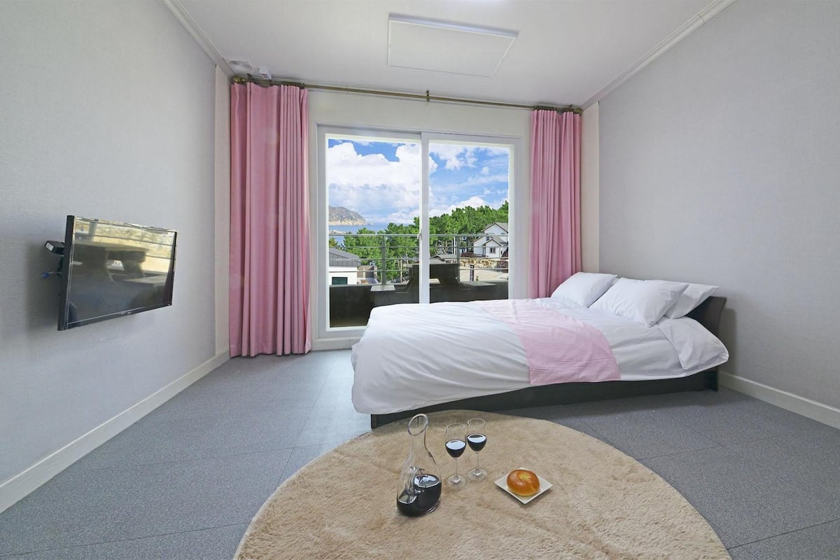 Une chambre à coucher avec un lit, un téléviseur fixé au mur, une fenêtre avec des rideaux et un tapis rond sur lequel se trouvent une bouteille et des verres.