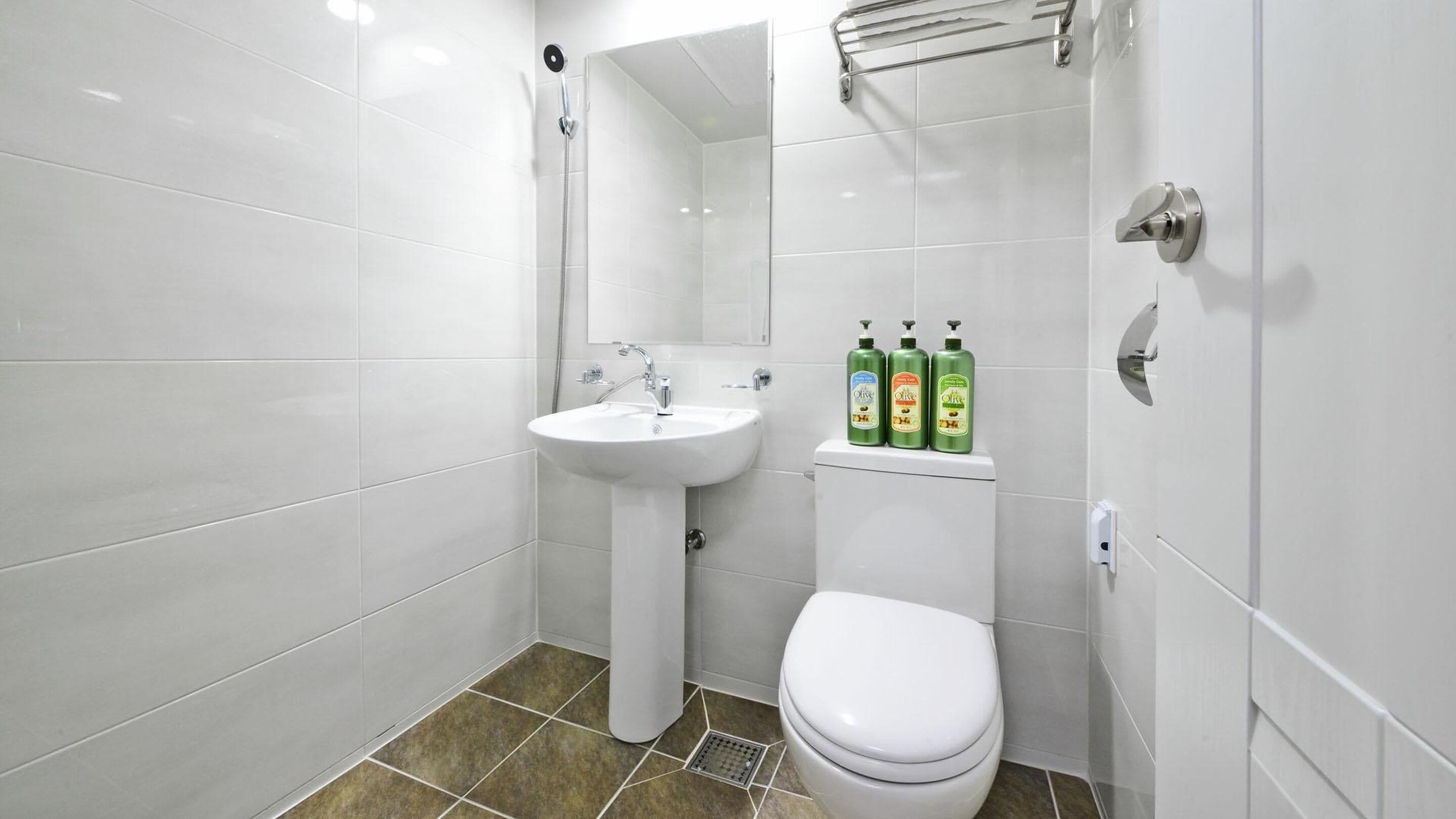 Un baño moderno con azulejos blancos, un lavamanos pedestal, un inodoro y tres botellas de aceite de oliva en un estante.