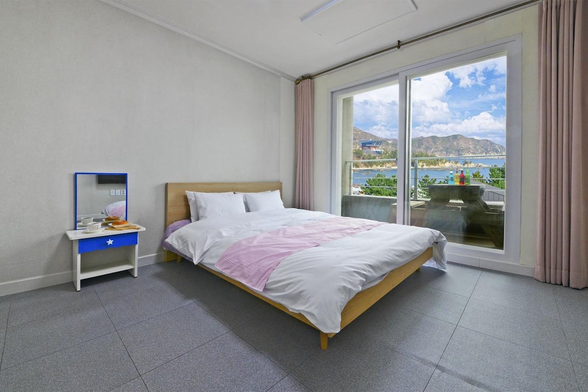 Une chambre d’hôtel avec un lit, un bureau doté d’un miroir et une grande fenêtre donnant sur la mer et les montagnes.