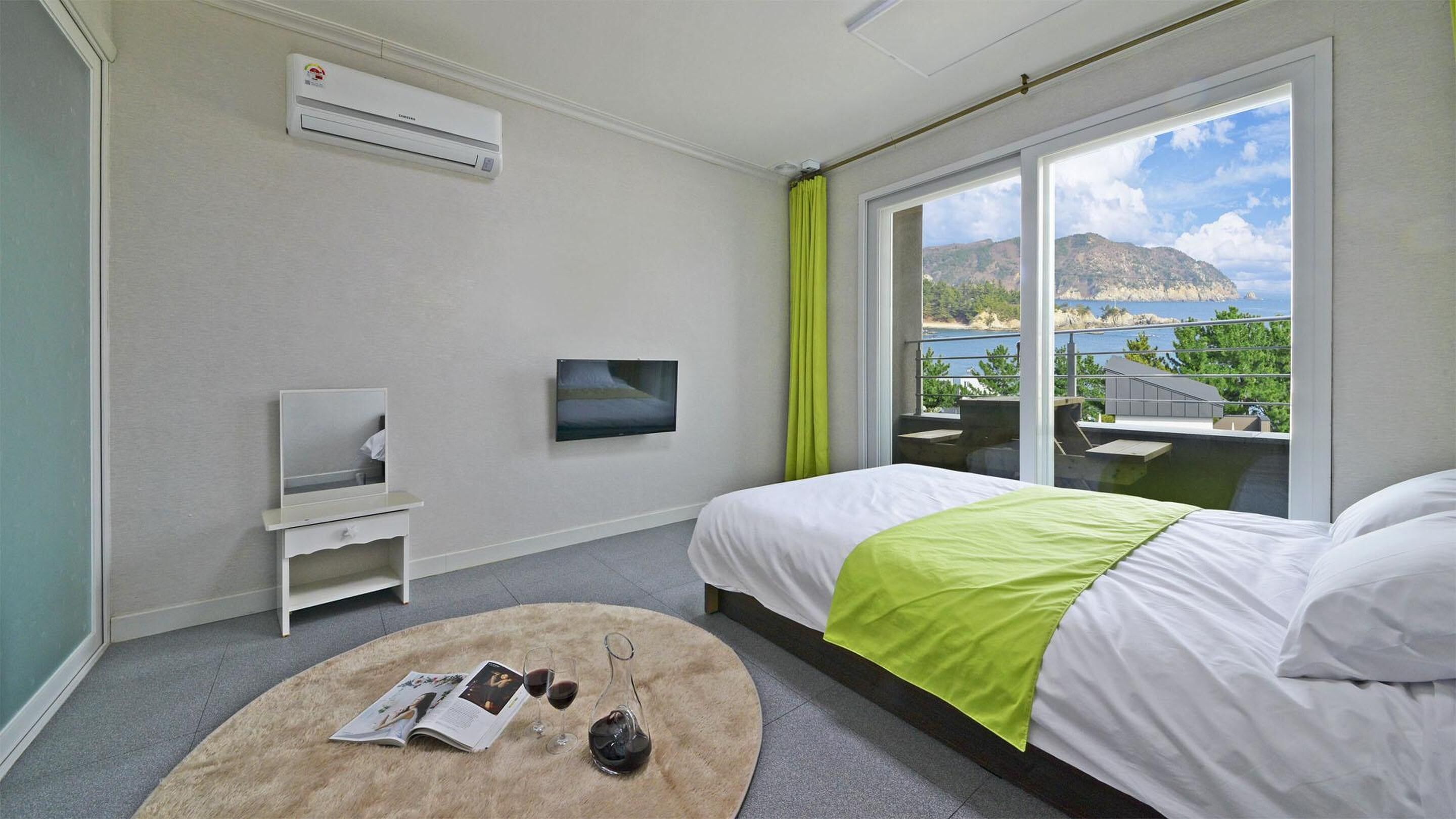 A hotel room with a bed, a TV, a window with a view of the sea, and a small table with a magazine and glasses.